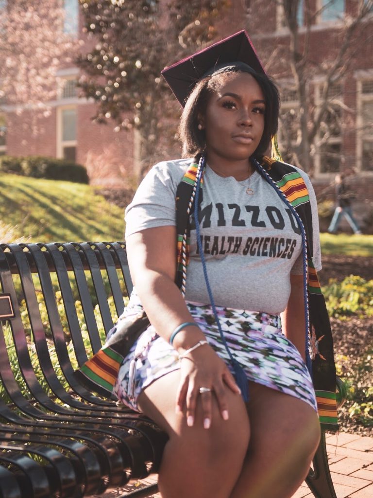 Aliyha Hill, sitting on a bench on Mizzou's campus in spring or summer, wearing a Mizzou Health Sciences grey T shirt with a floral skirt. She's also wearing her graduation cap and cords