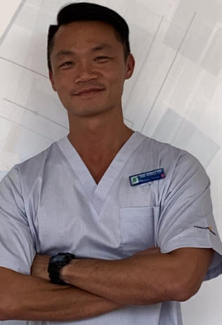 Ivan Lee, a respiratory therapist, standing in front of a white and grey back drop, wearing scrubs, with his arms crossed, in the hospital he works in
