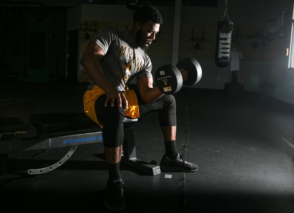 A man sits in a dramatically lit room lifting a large dumbell