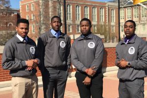 Four Black men with serious expressions wearing matching gray quarter-zip jackets