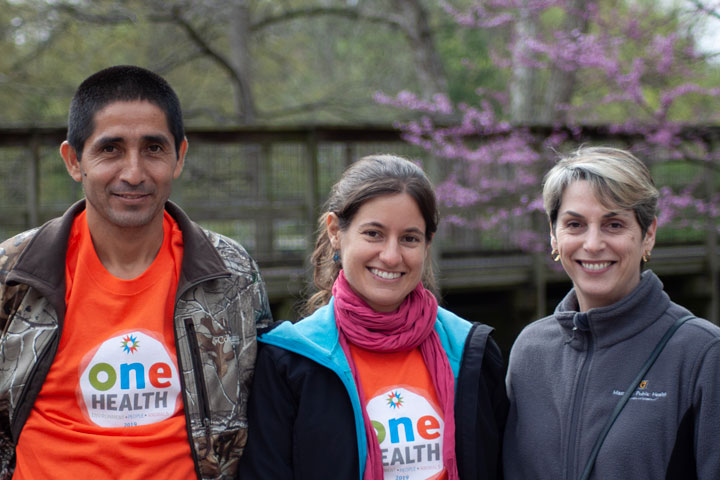 Two students wearing orange "One Health" T-shirts pose for a photo with Public Health professor Lisa Saffran