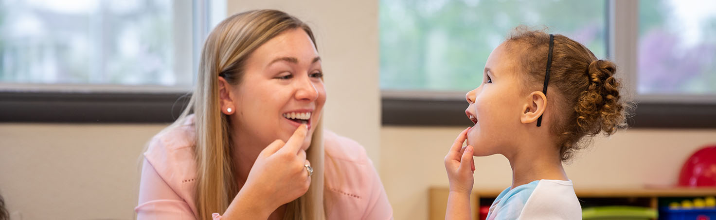 Student points to her mouth while giving therapy services to a child