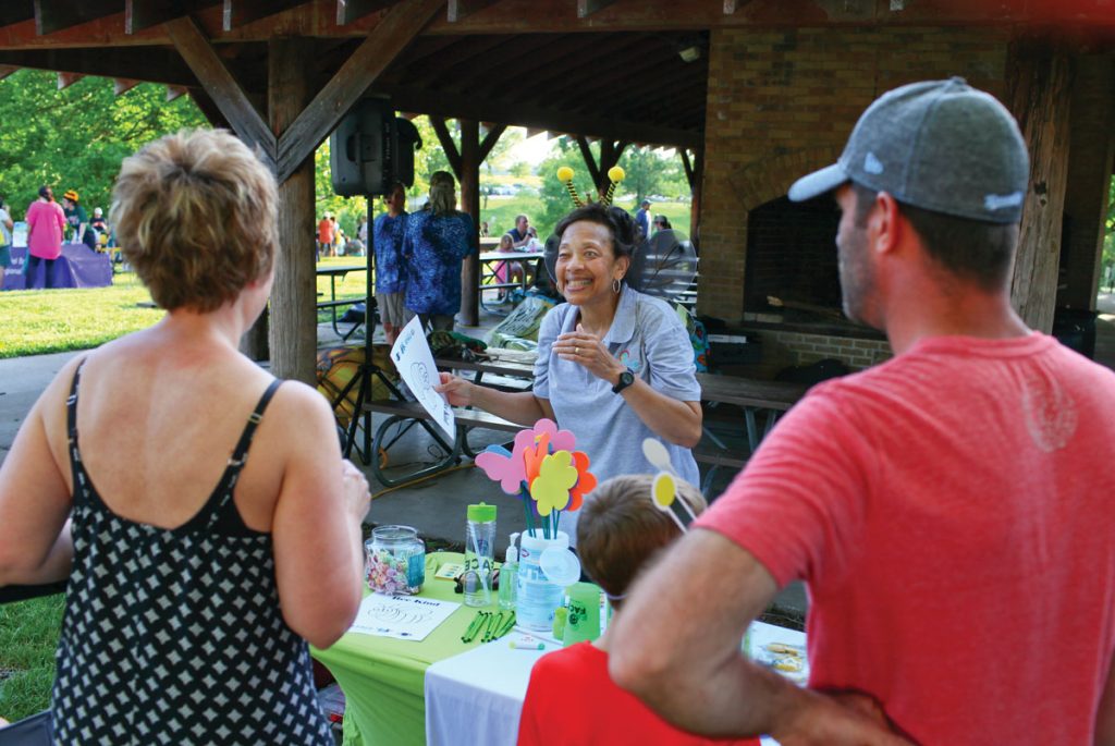 A FACE staff member engages with community members across a brightly decorated table at a park pavilion.