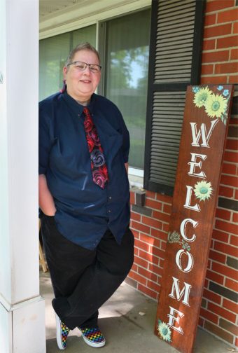 A woman in a button down shirt and red paisley tie stands next to a wooden sign that says "Welcome"