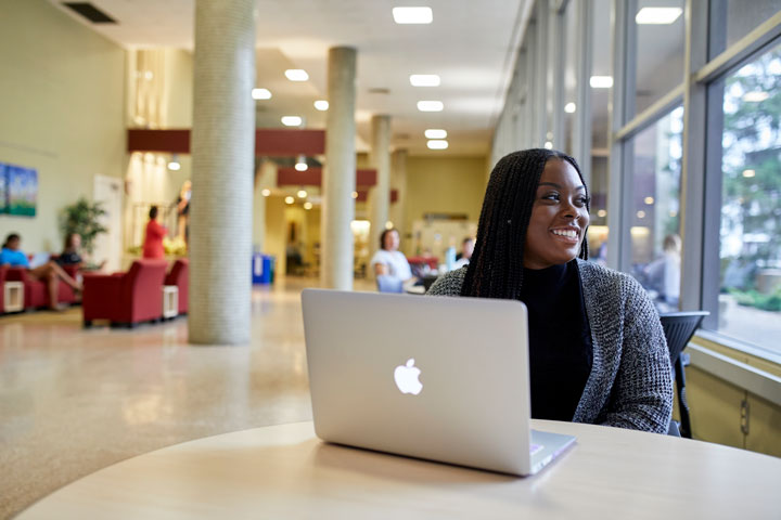 A student sits in the lobby in front of a laptop. She is looking out the window and smiling slightly.