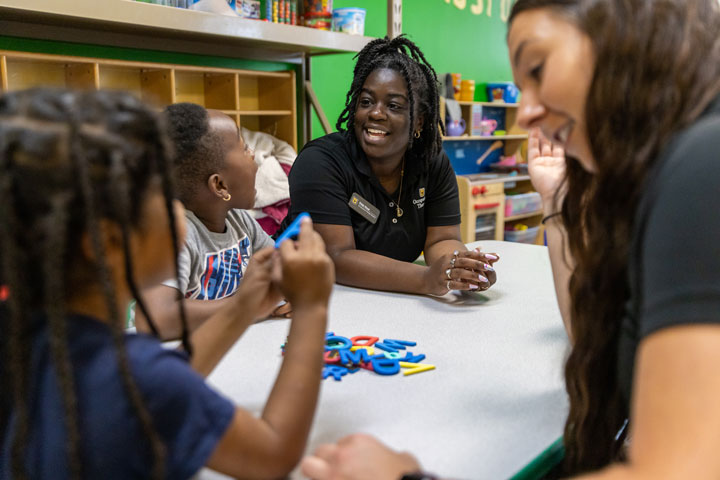 College students sit around a table with preschool-aged children in a colorful room