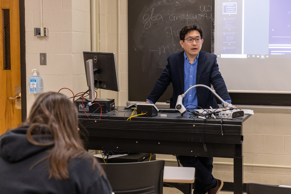 Professor leans on desk while speaking to class