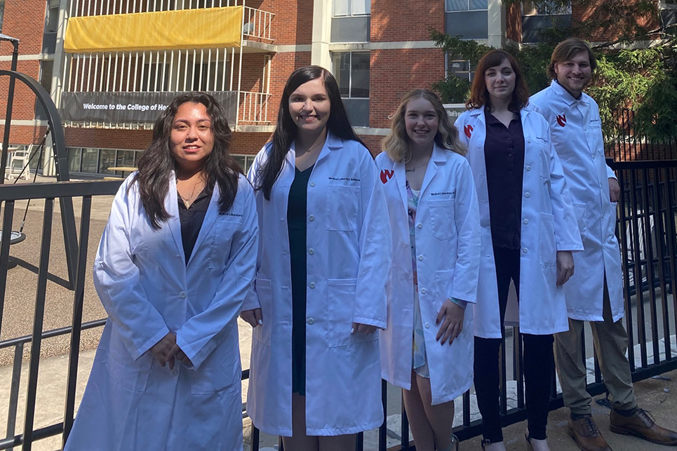 Students pose in a courtyard wearing white laboratory coats.
