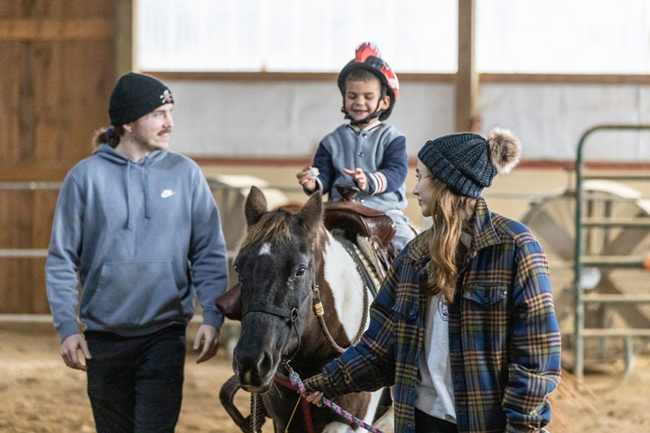 two college students help guide a horse with a child sitting on it