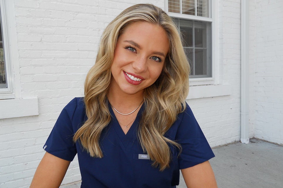 A young woman in scrubs stands outside of a white brick building