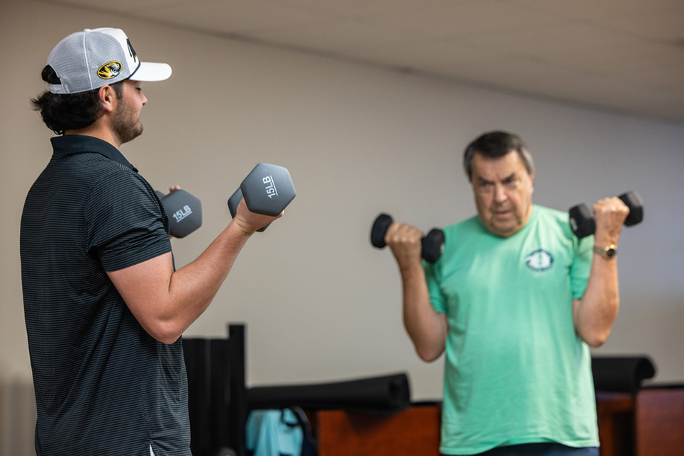 A young man coaches an older man in how to curl dumbbells