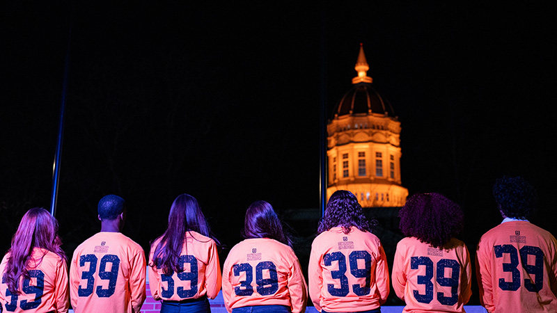 Students in shirts labeled with 39 face Jesse Hall during a ceremony