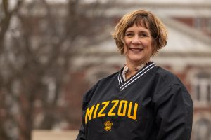 A woman stands outside of a building while wearing a black Mizzou jacket