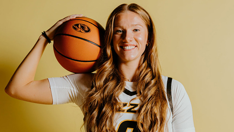 A young woman holds a basketball on her shoulder